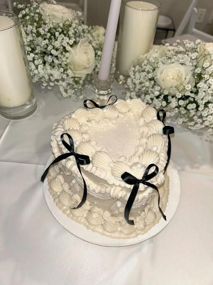 Heart-shaped vintage white cake with intricate buttercream piping, white pearls, and black satin bows on a white base.