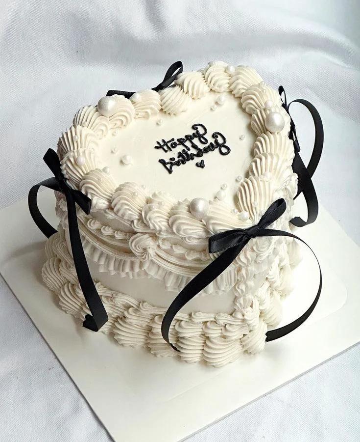Heart-shaped white vintage cake with intricate ruffles, black satin bows, and pearl accents for a birthday celebration.
