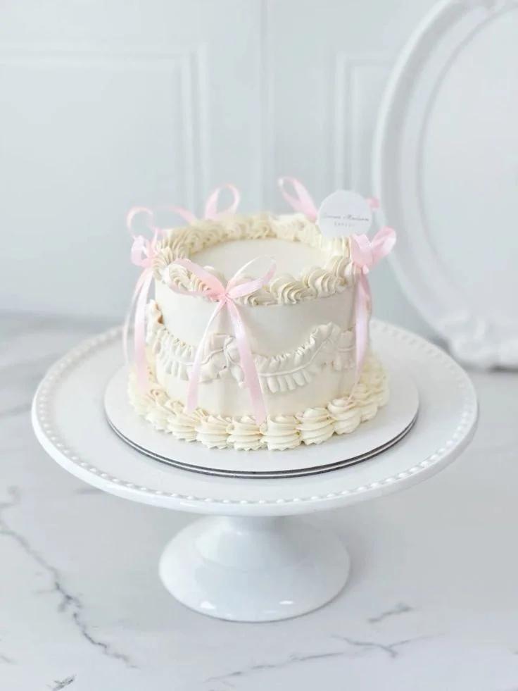 Elegant white vintage-style cake with pink satin ribbons and intricate piped ruffles on a white pedestal stand.