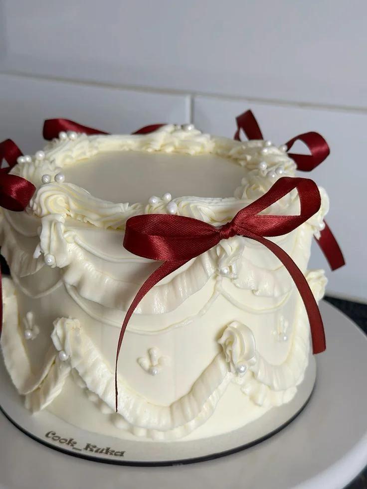 Elegant white vintage-style cake with intricate buttercream ruffles, white pearls, and deep red satin bows.