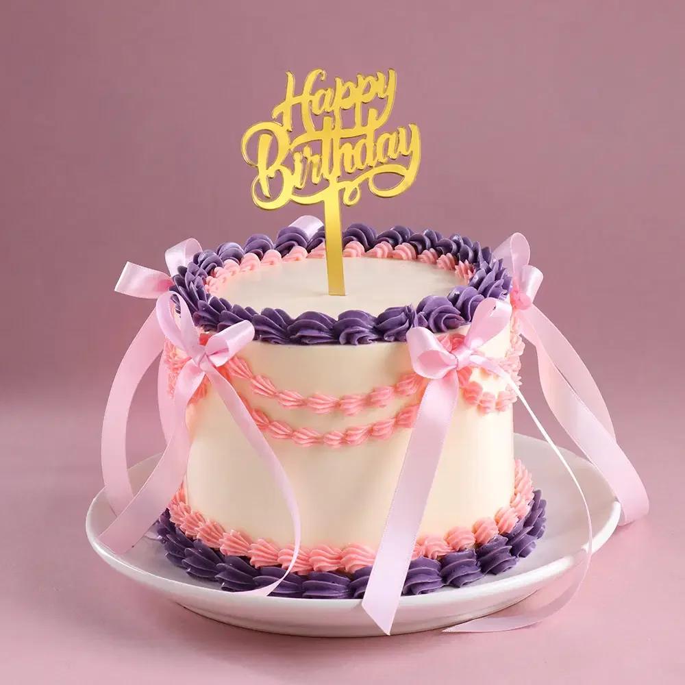 Elegant vintage-style white cake with purple and pink piped icing borders, pink satin ribbons, and a gold Happy Birthday topper.