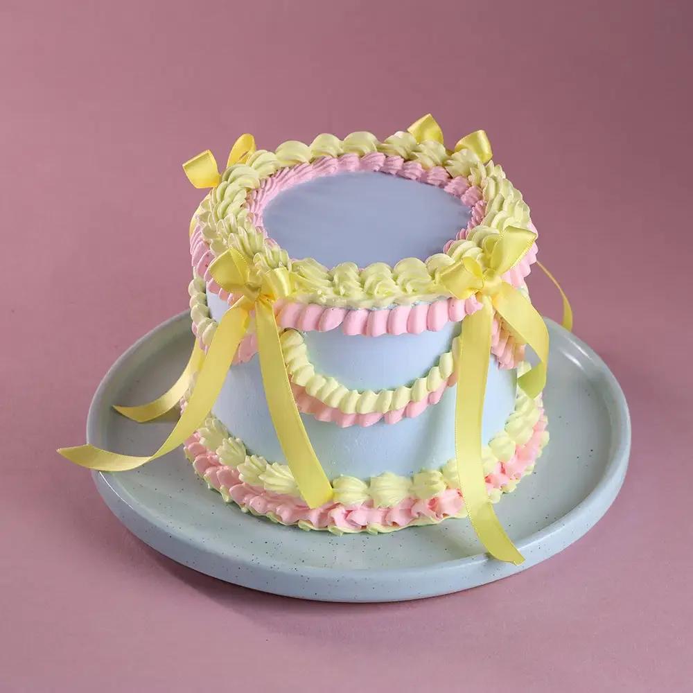 A tall light blue vintage-style cake with intricate pink and light yellow buttercream ruffles and yellow satin ribbons.