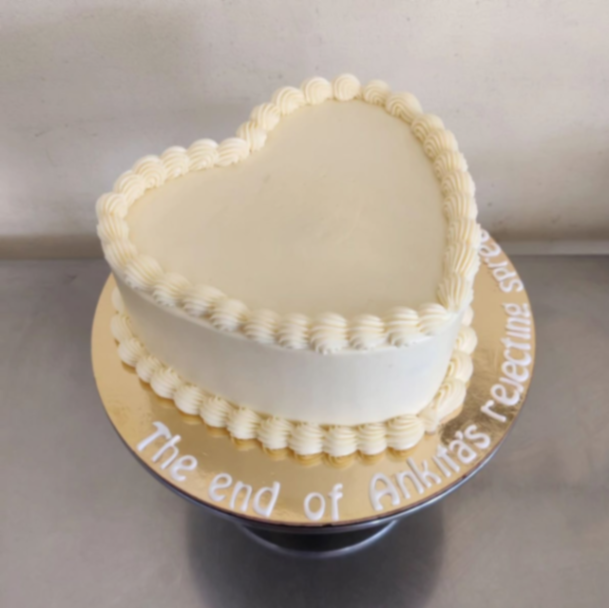 White heart-shaped minimalist cake with piped icing borders and a message on a gold base board.