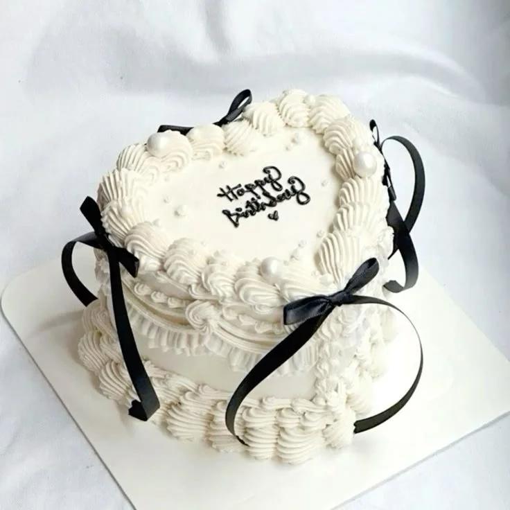 White heart-shaped vintage cake with intricate piped ruffles, black satin bows, and white pearls featuring a Happy Birthday message.