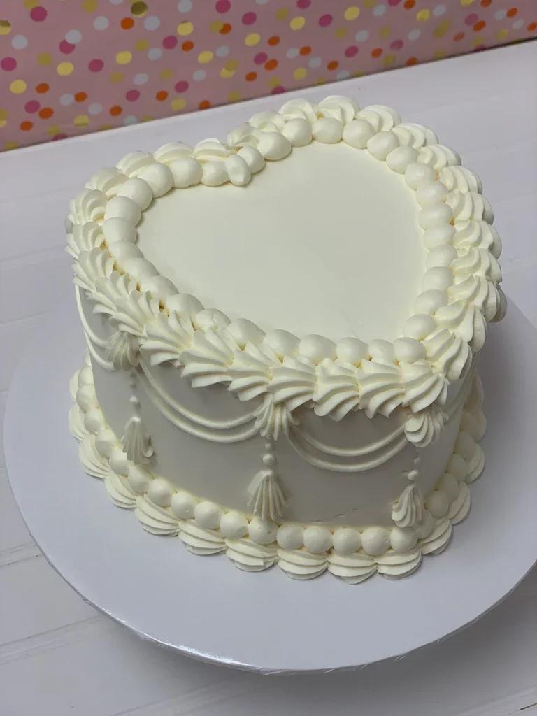 White heart-shaped vintage cake with intricate buttercream piping, ruffles, and tassels on a white base.