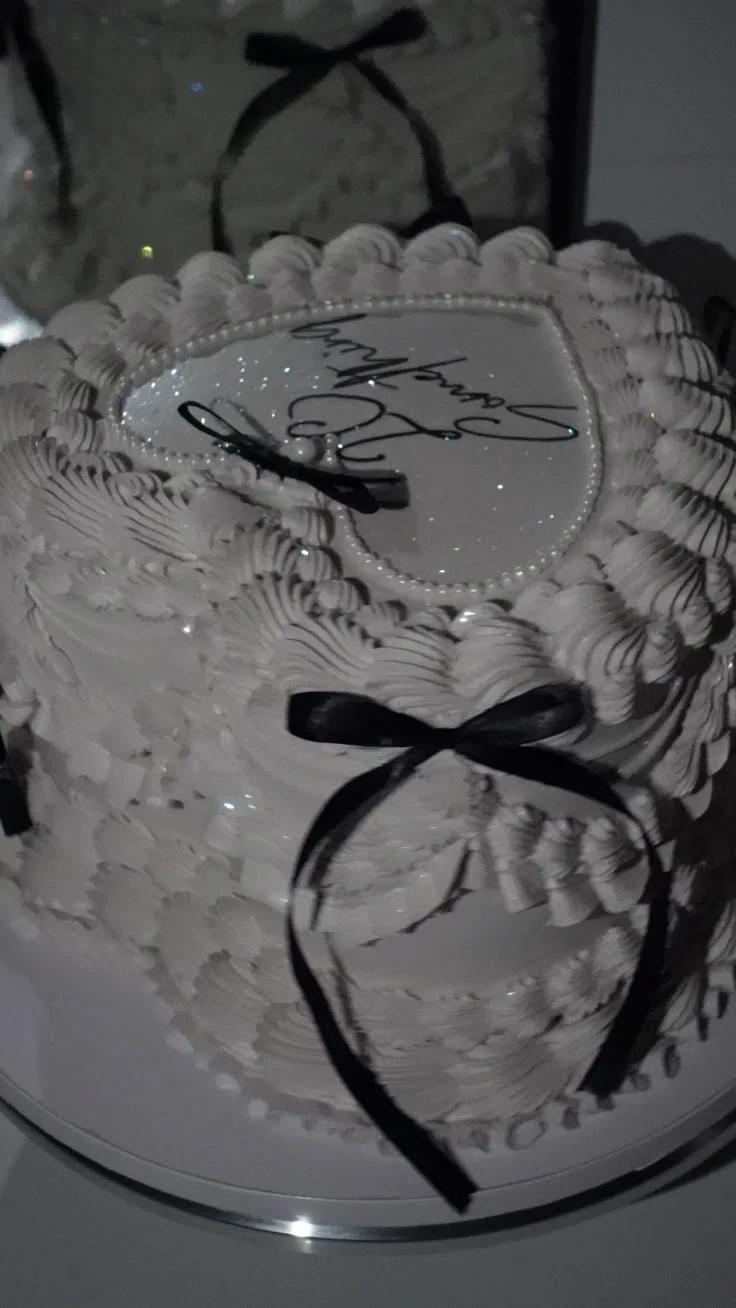 White heart-shaped vintage cake with intricate piped ruffles, black satin bows, and a personalized message on top.