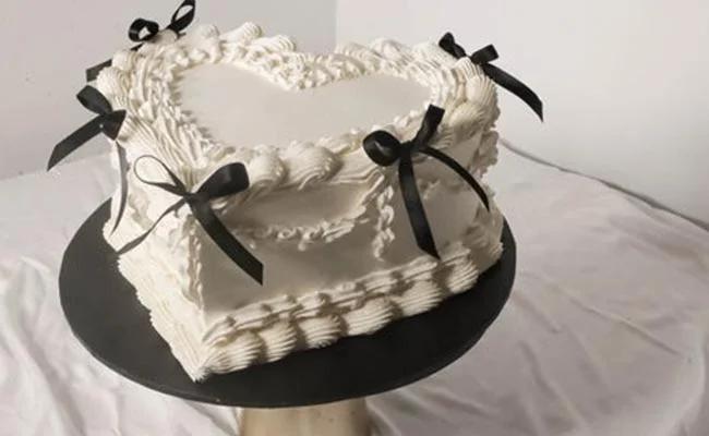 Heart-shaped white vintage cake with intricate piped ruffles and black satin bows on a black cake stand.